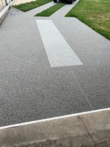 Grauer Steinteppich als fugenlose Oberflaeche auf Einfahrt und Gehweg mit hellem Designstreifen vor dem Haus